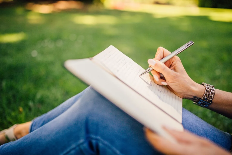 Henkilö istuu nurmella ja kirjoittaa jotain kynällä vihkoon. Henkilöllä on farkut jalassa ja kello kädessä.