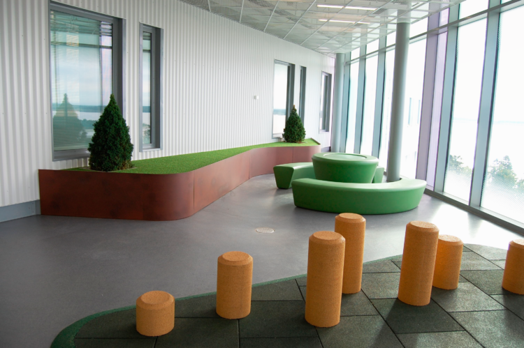 Bilden visar ett podium med två gröna växter ovanpå, ett grönt runt bord bredvid och gröna bänkar svängda runt. I förgrunden gula stolpar på grön bas, som man kan klättra på.