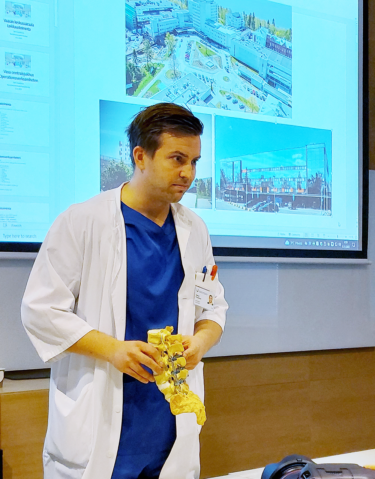 En läkare med en blå skjorta och en vit jacka håller i sina händer en tredimensionell modell av ryggraden, som illustrerar skruvarna och metallstöden från förbeningsoperationen.