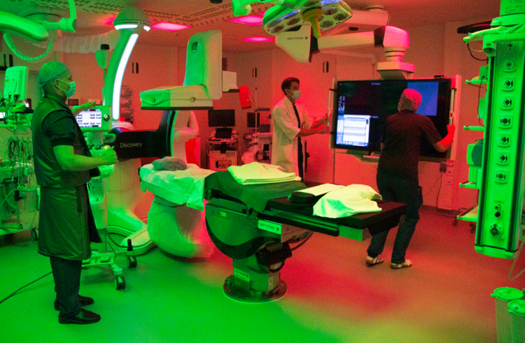 Leikkaussalin keskellä on tyhjä hoitotaso, vasemmalla seisoo kirurgi sädesuojavarustuksessa, oikealla lääkäri ja hoitaja siirtävät isoa näyttöruutua lähemmäs hoitotasoa. Huoneen vasen puoli on valaistu vihreällä valolla ja oikea puoli punaisella valolla.