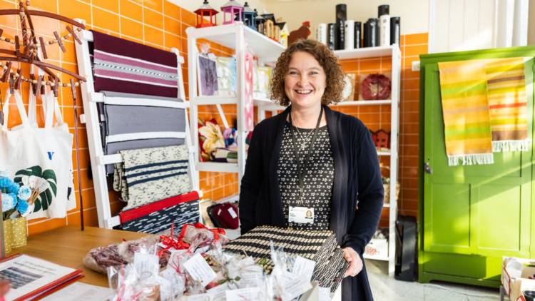 Sonja Tallgård står i Regnbågens butik bland mattor, tändrosor, tvålar och andra produkter som är till salu. Hon har brunt, lockigt hår och är klädd i en svart kofta.