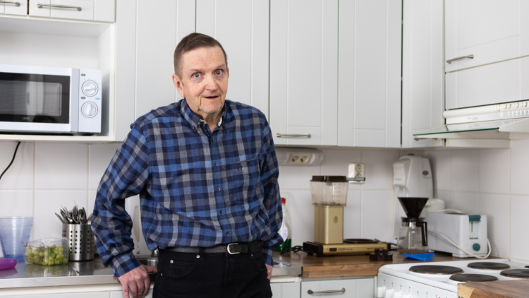 Carl-Erik Sundqvist står i sitt kök och lutar sig mot ett köksskåp. Han är klädd i en blårutig kragskjorta och mörka byxor.