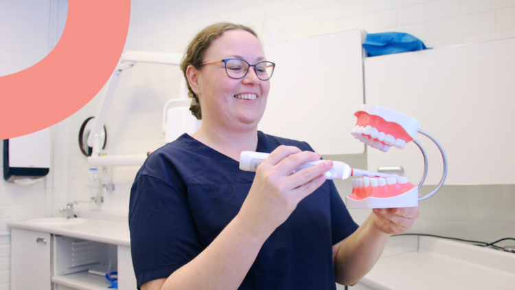 Pia Lindbäck seisoo hammashoitohuoneessa ja pitelee käsissään isoja muovisia tekohampaita, joita hän harjaa sähköhammasharjalla.