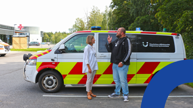 Nainen ja mies seisovat ambulanssinnäköisen auton vierellä keskustelemassa. Mies osoittaa auton katolla olevaa sinistä vilkkuvaloa.