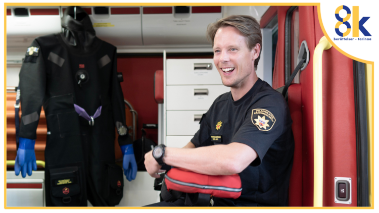En leende brandman iklädd kortärmad, svart skjoita med räddningsverkets emblem sitter i baktill i en brandbil. Till höger syns en dykardräkt.