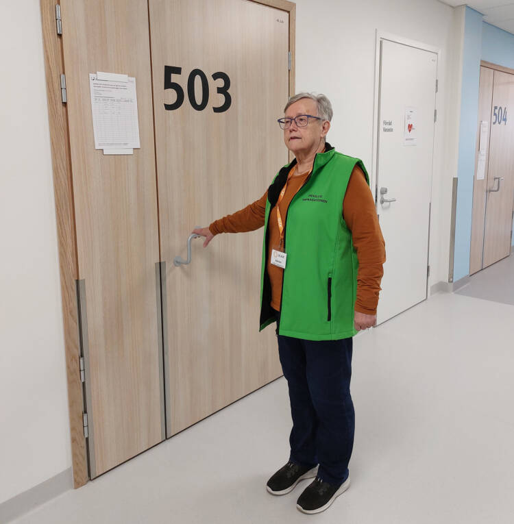 Nainen, jolla on vihreä liivi yllään seisoo sairaalan käytävällä ja tarttuu potilashuoneen ovenkahvaan.