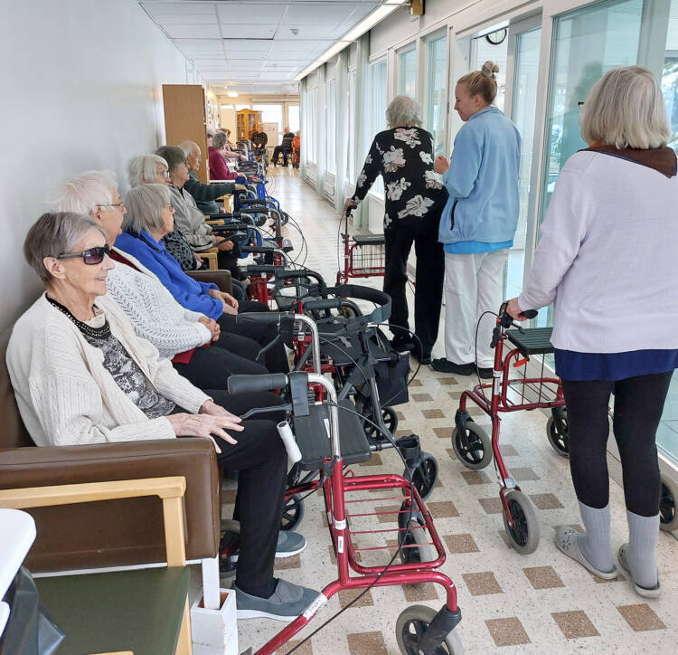 Käytävän seinustalla on rivissä tuoleja, joissa istuu kymmeniä ikäihmisiä, jokaisella on edessään rollaatori.