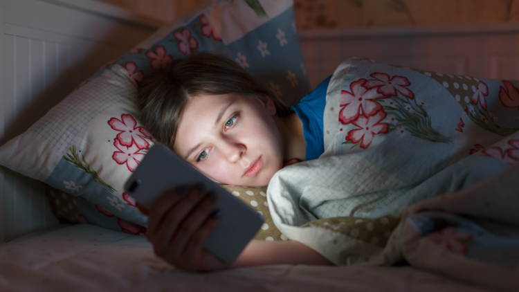 En ung person ligger i säng och kollar sin mobiltelefon.